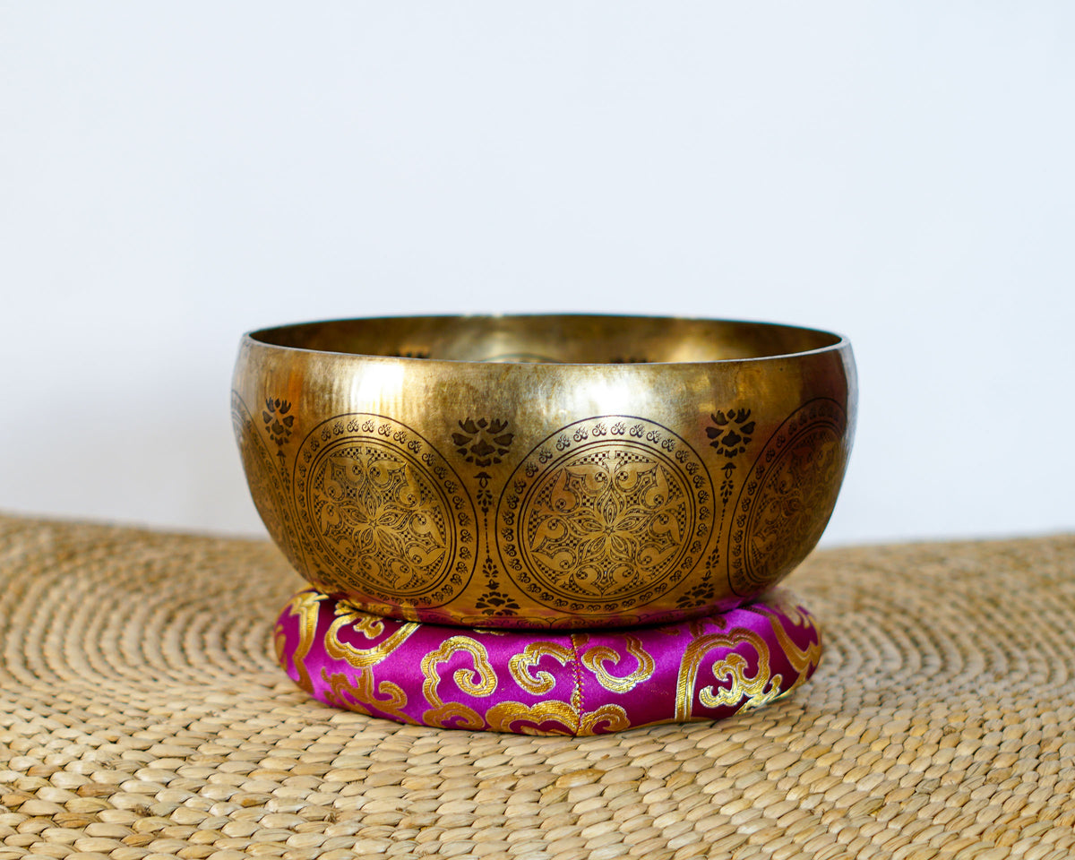 Golden Avalokiteshvara singing bowl with decorative circular engravings displayed on a purple brocade cushion.