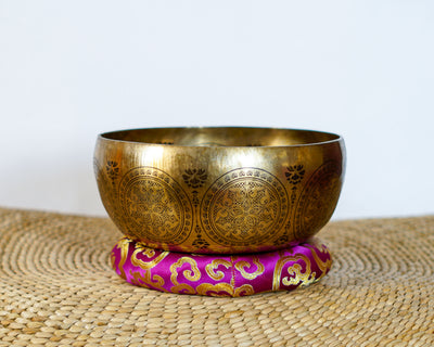 Golden Avalokiteshvara singing bowl with decorative circular engravings displayed on a purple brocade cushion.