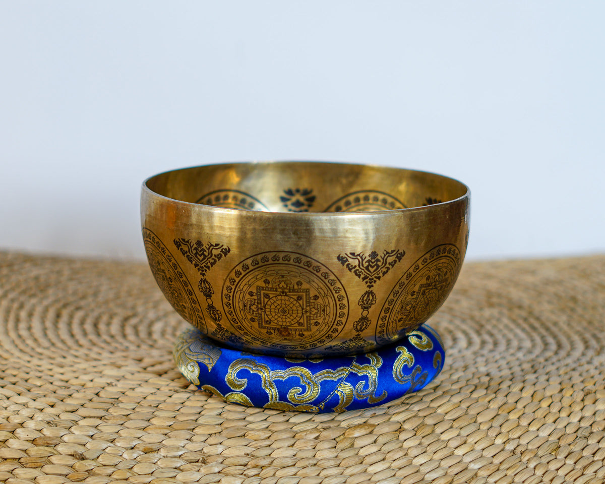 Seven Chakra Singing Bowl resting on a blue brocade cushion, featuring intricate mandala engravings on the exterior.