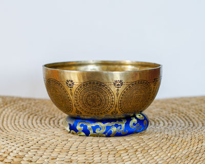 Kalchakra Yogini Singing Bowl resting on a blue brocade cushion, displaying intricate mandala engravings on its exterior.