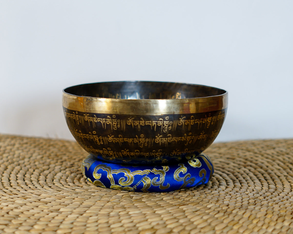 Dark golden Om Mantra singing bowl with repeated Tibetan mantra inscriptions around its exterior, displayed on a blue brocade cushion.