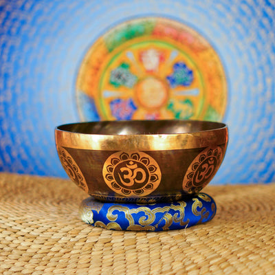 Handcrafted Om singing bowl from Nepal with golden Om engravings, dark bronze finish, decorative blue cushion and wooden striker.