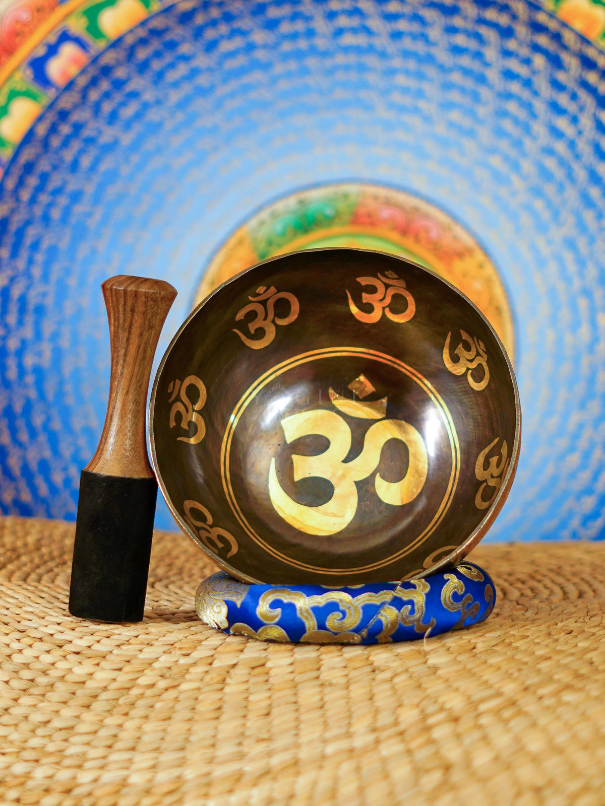 Handcrafted Om singing bowl from Nepal with golden Om engravings, dark bronze finish, decorative blue cushion and wooden striker.