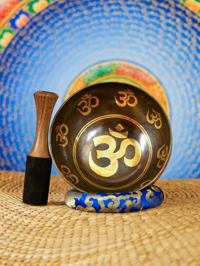 Handcrafted Om singing bowl from Nepal with golden Om engravings, dark bronze finish, decorative blue cushion and wooden striker.