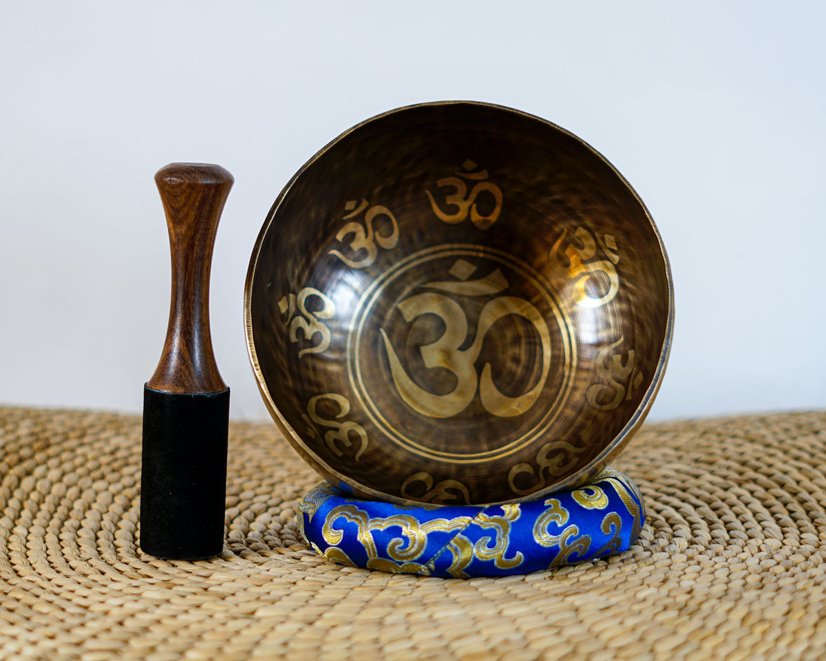 Hindu Om engraved singing bowl displayed upright on a blue brocade cushion beside a wooden mallet, showing the interior Om symbol and decorative borders.