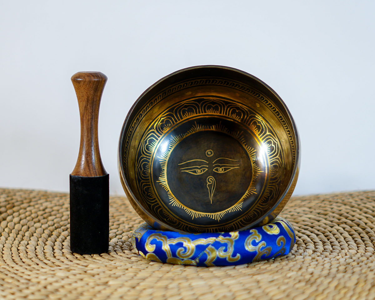 Dark antique gold Buddha Eye singing bowl with golden Tibetan inspired decorative motifs on a blue brocade cushion.