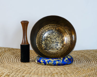 Tree of Life engraved singing bowl displayed upright with a wooden mallet and blue brocade cushion on a woven mat.