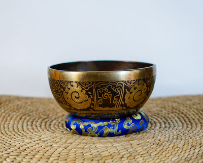 Ganesh engraved metal singing bowl with antique gold detailing and a blue brocade cushion on a woven mat.