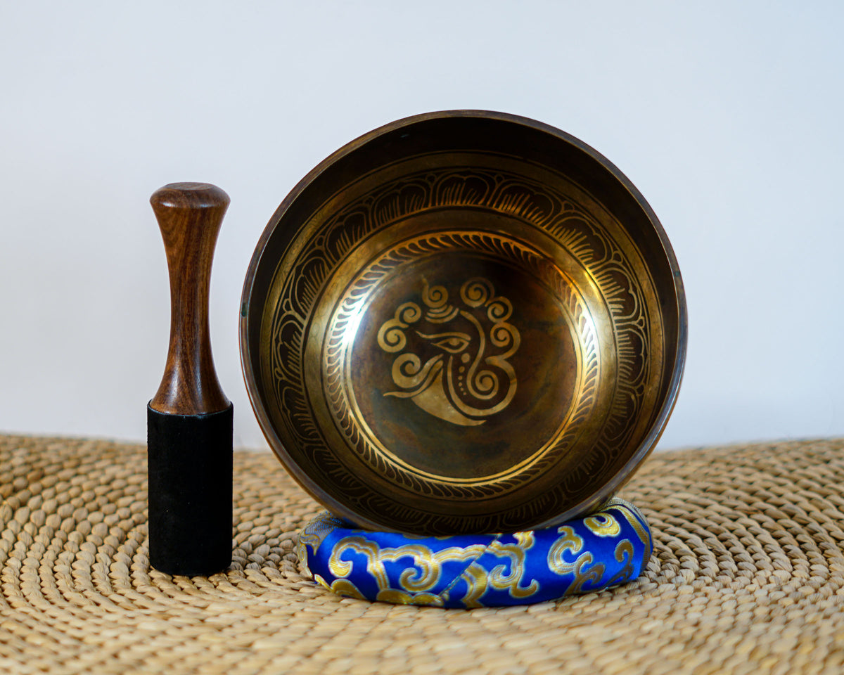 Ganesh singing bowl displayed upright with a wooden mallet and blue brocade cushion, showcasing its detailed interior engraving.