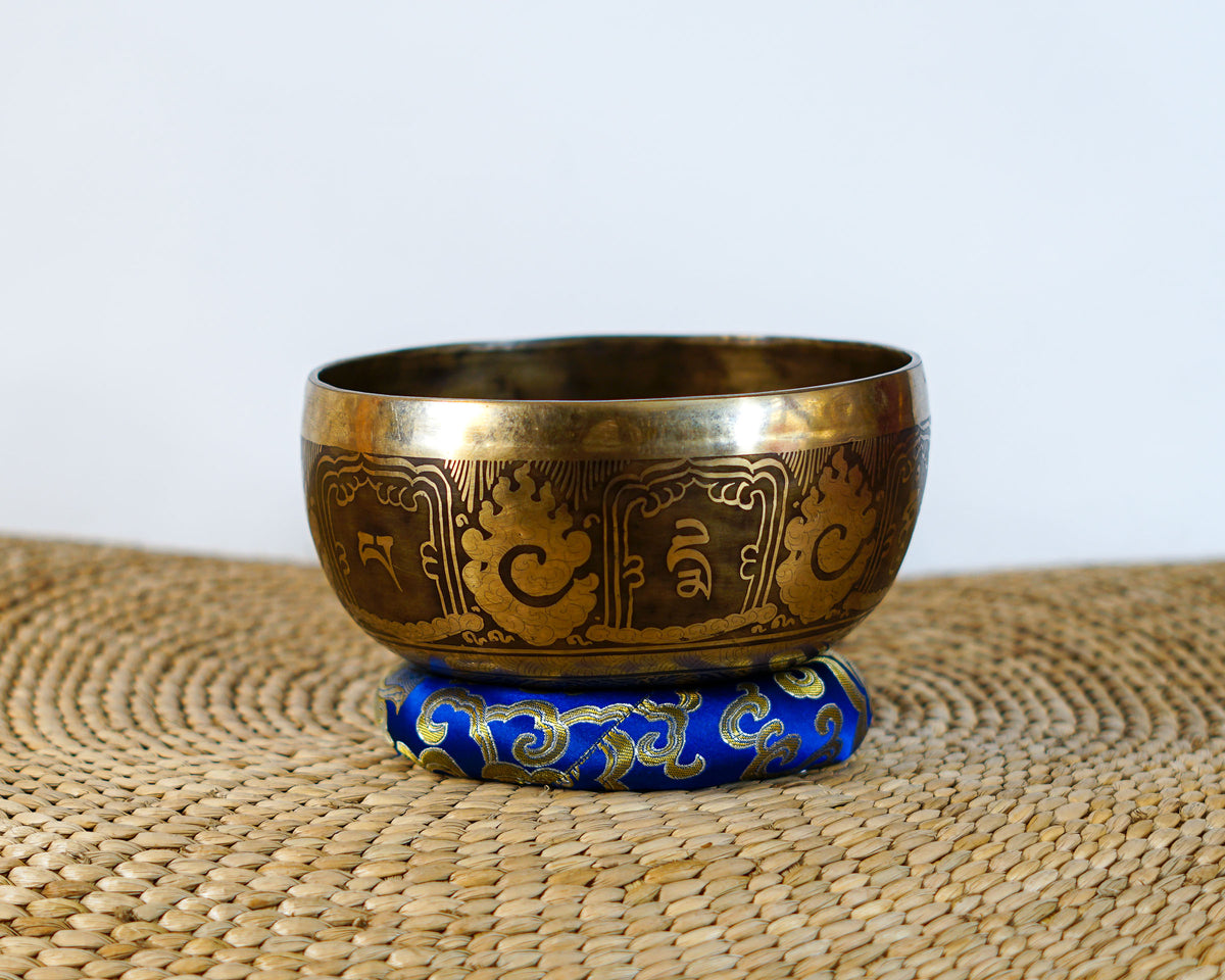 Hindu Om engraved singing bowl with dark decorative patterns and a blue brocade cushion on a woven mat.