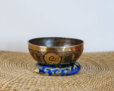 Hindu Om singing bowl with antique gold and dark engraved exterior displayed on a blue brocade cushion.