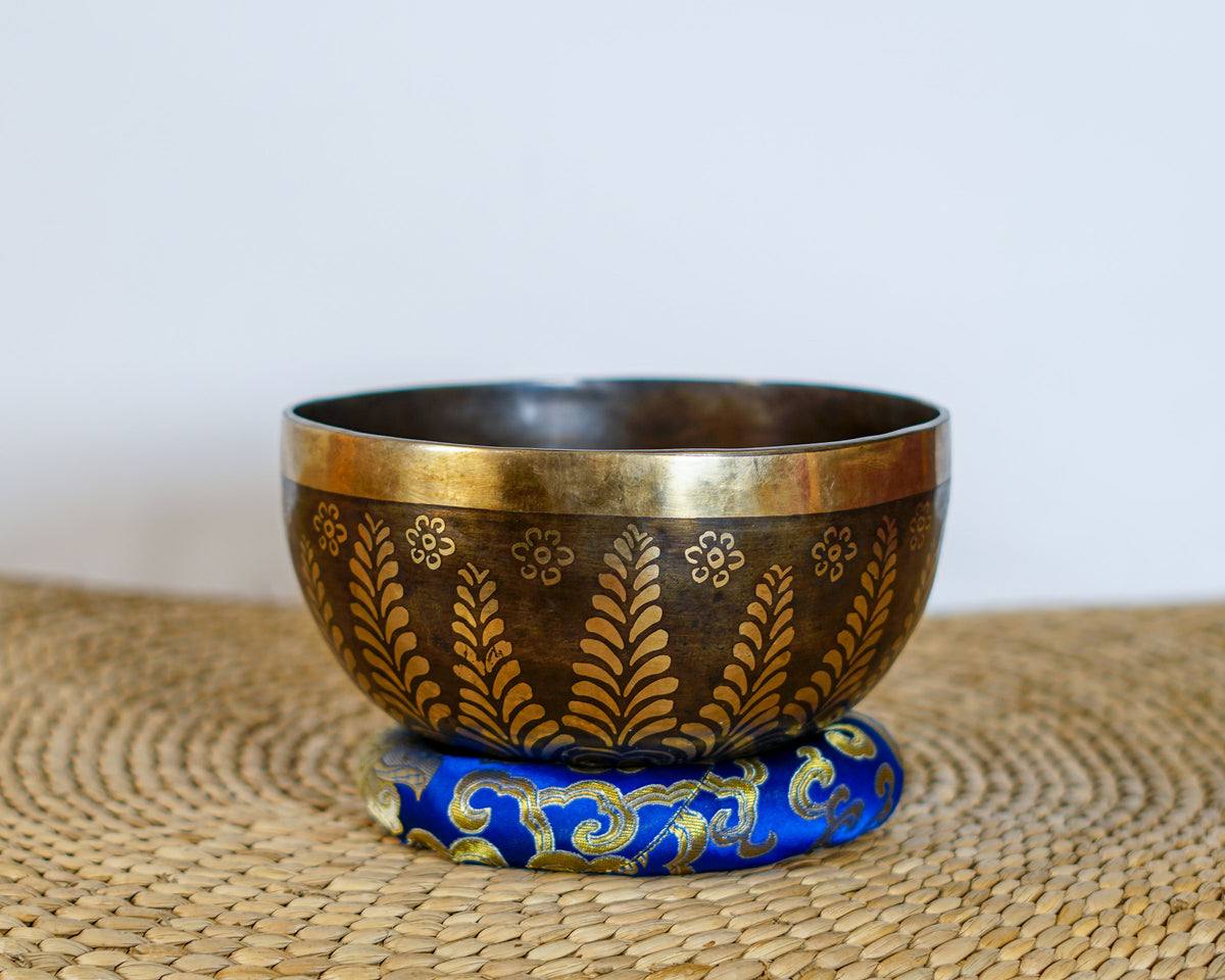 Flower Yin Yang singing bowl with gold engraved floral motifs and a dark antique finish displayed on a blue brocade cushion.