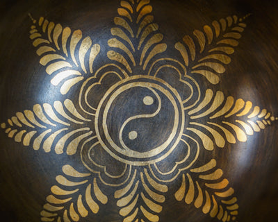 Close up of a golden Yin Yang symbol surrounded by a decorative floral pattern inside a dark metal singing bowl.