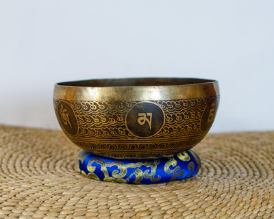 Lotus engraved singing bowl with golden exterior patterns resting on a blue brocade cushion.
