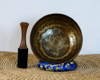 Engraved lotus singing bowl displayed upright with a wooden mallet and blue brocade cushion, showing the intricate golden lotus design inside.