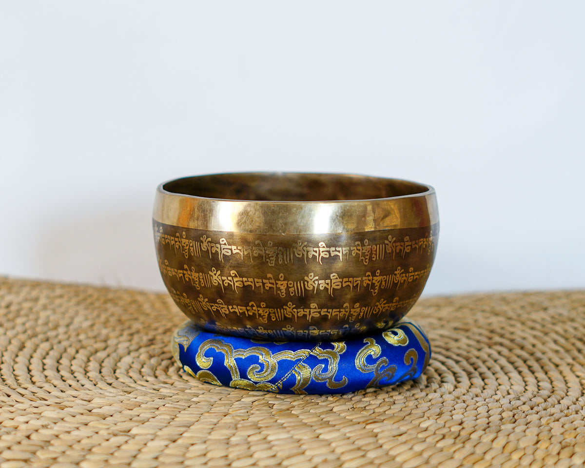 Flower of Life engraved singing bowl with Tibetan lettering around the exterior, displayed on a blue brocade cushion.