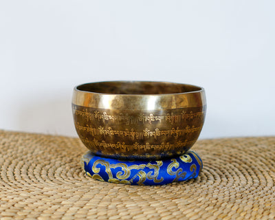 Flower of Life engraved singing bowl with Tibetan lettering around the exterior, displayed on a blue brocade cushion.