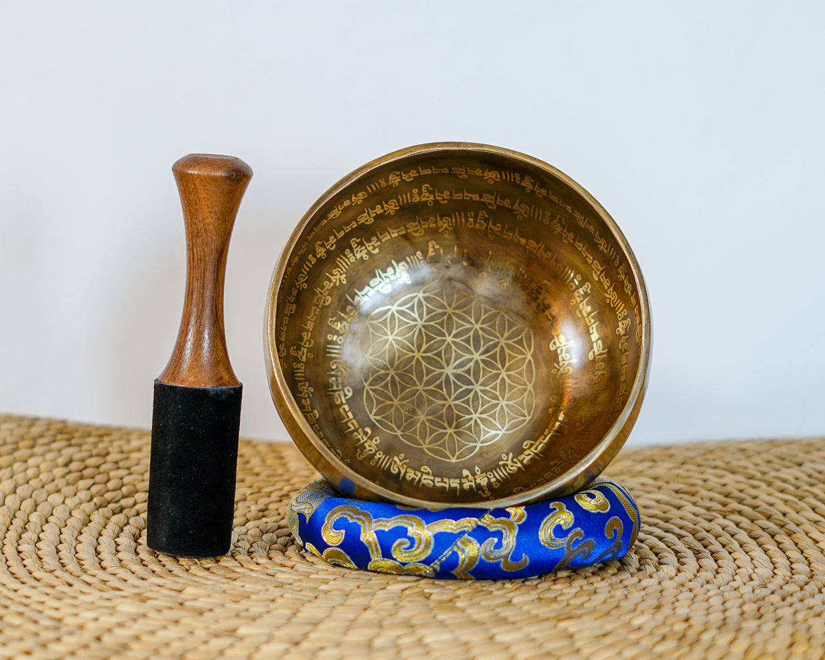 Singing bowl displayed upright with a wooden mallet and blue brocade cushion, showing the central Flower of Life geometric pattern and Tibetan script.
