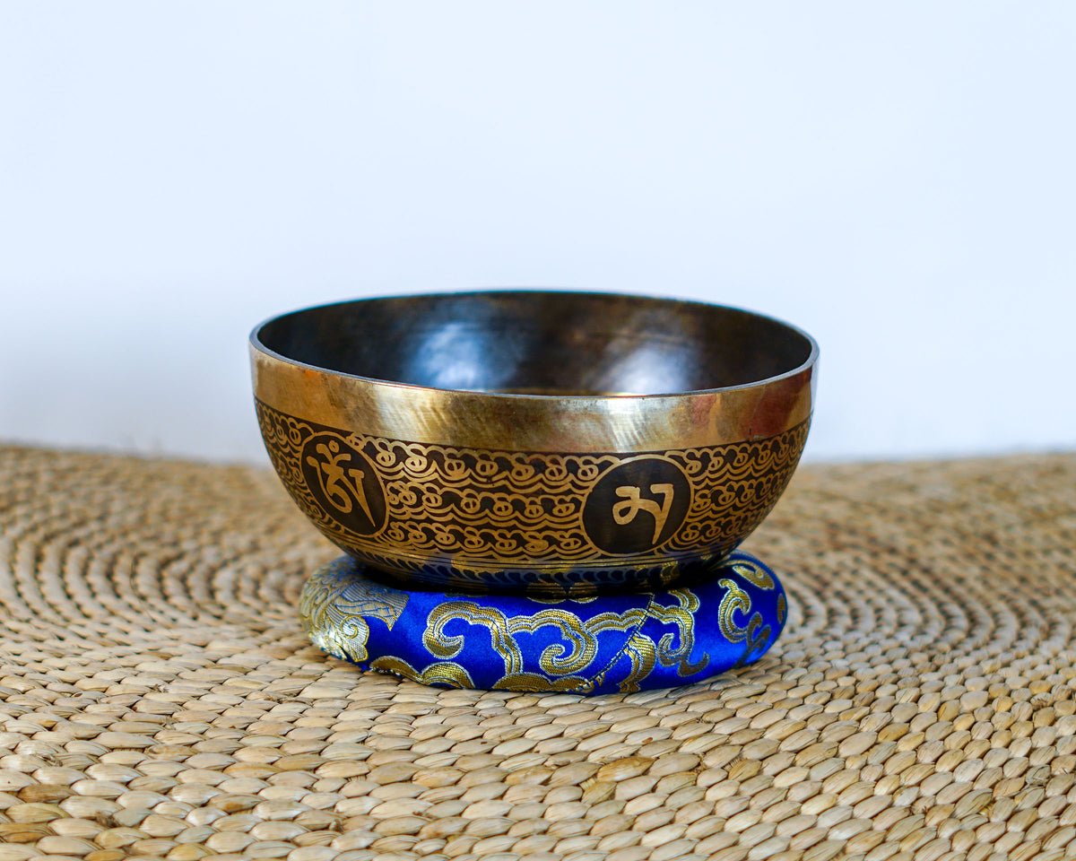 Antique gold singing bowl featuring Tibetan script and intricate decorative patterns on a blue brocade cushion.