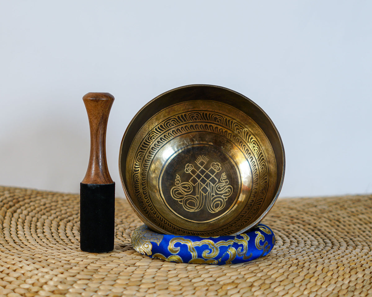 Engraved Endless Knot singing bowl displayed upright on a blue brocade cushion beside a wooden mallet, showing the central symbol and ornate interior borders.
