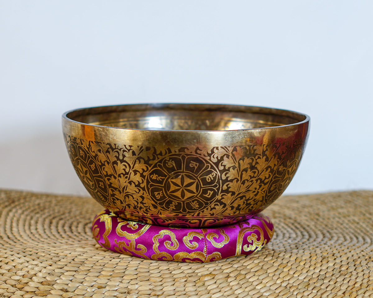 Golden and dark bronze Chakra Manab Tibetan singing bowl with intricate floral and geometric engravings displayed on a pink brocade cushion.