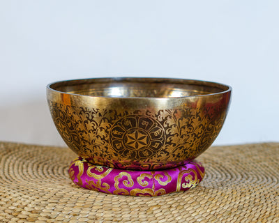 Golden and dark bronze Chakra Manab Tibetan singing bowl with intricate floral and geometric engravings displayed on a pink brocade cushion.