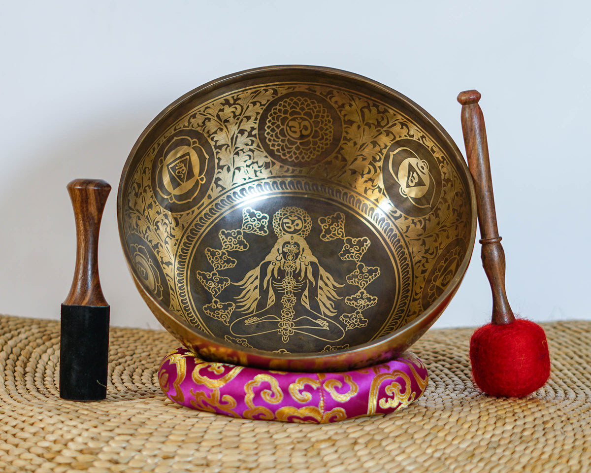 Chakra Manab engraved singing bowl shown upright with a meditating figure design, decorative borders, brocade cushion, and wooden strikers.