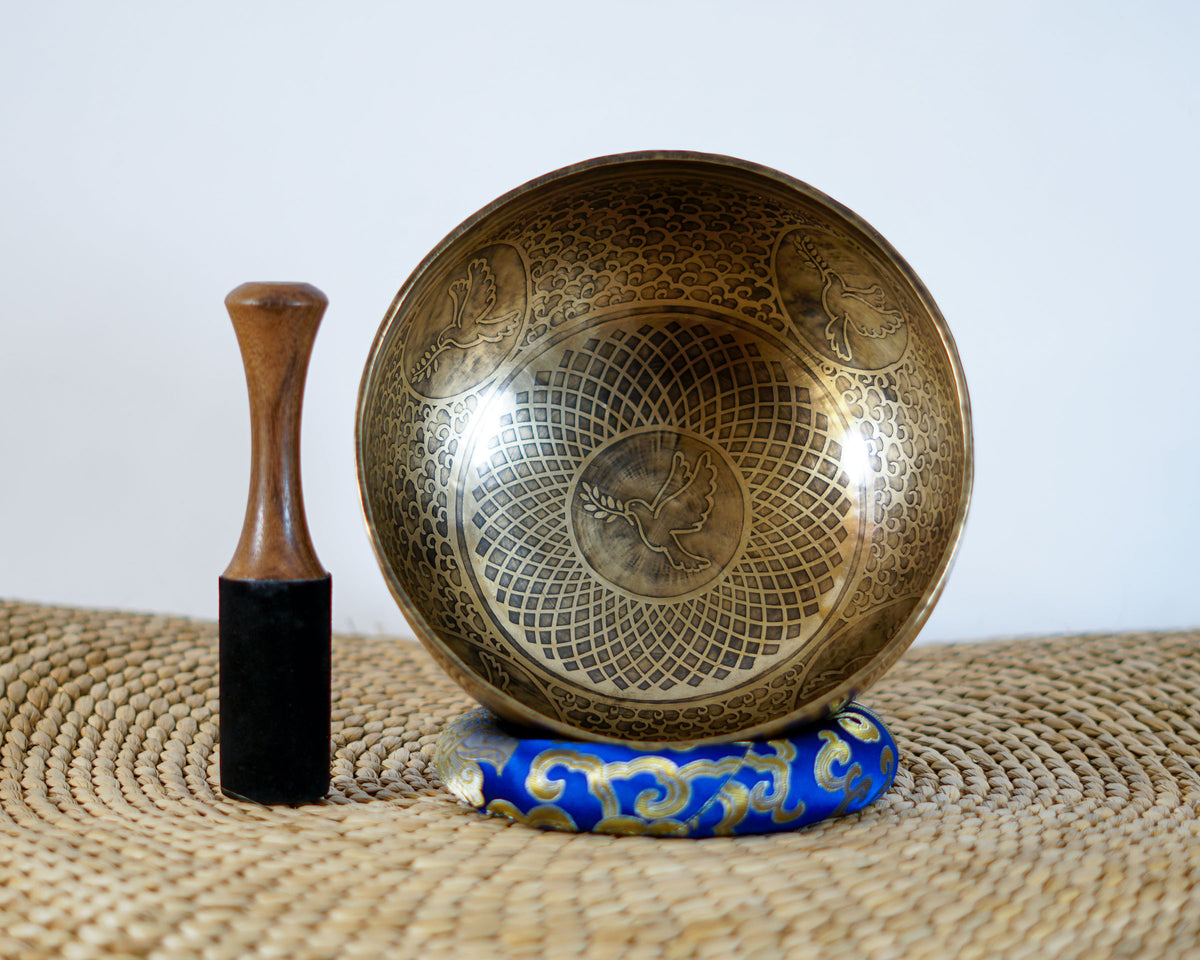Dove engraved Tibetan singing bowl displayed upright with intricate geometric patterns, a blue brocade cushion, and a wooden striker.