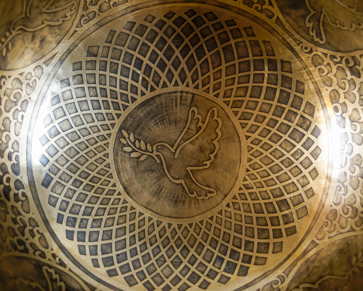 Close up of a Tibetan singing bowl engraved with a dove carrying an olive branch, surrounded by geometric lattice patterns and decorative motifs.