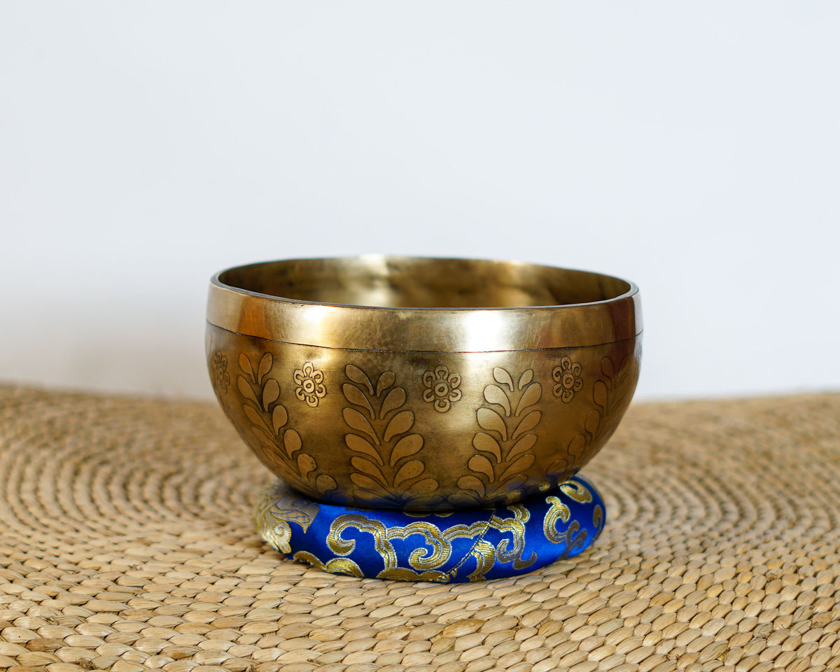 Antique golden Tree of Life Tibetan singing bowl with engraved leaf and floral patterns displayed on a blue brocade cushion.