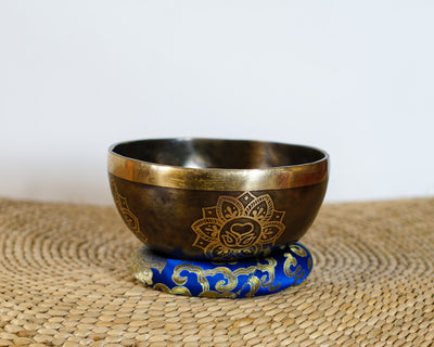 Dark antique bronze Tibetan singing bowl with a golden Swan Symbol of Life engraving on the exterior, displayed on a blue brocade cushion.