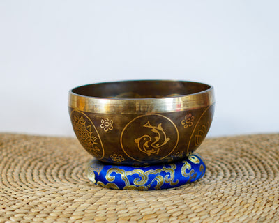 Side exterior view of a dark antique bronze Tibetan singing bowl with golden engraved fish motifs, resting on a blue brocade cushion.