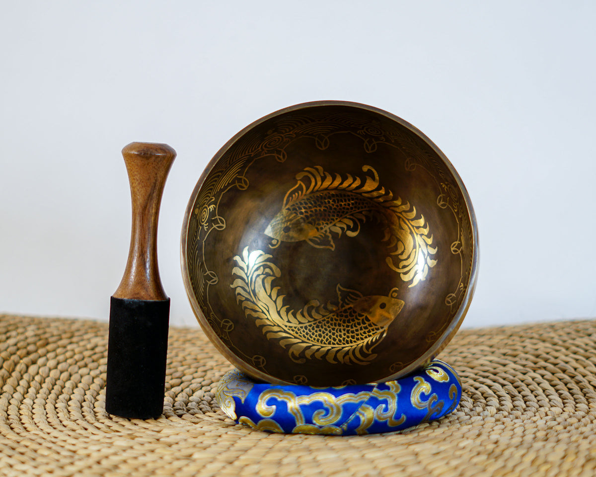 Angled top view of a dark bronze Tibetan singing bowl displaying a golden twin fish interior engraving, shown with a wooden mallet striker and blue cushion.