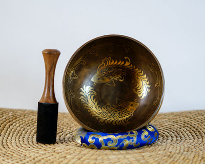 Angled top view of a dark bronze Tibetan singing bowl displaying a golden twin fish interior engraving, shown with a wooden mallet striker and blue cushion.
