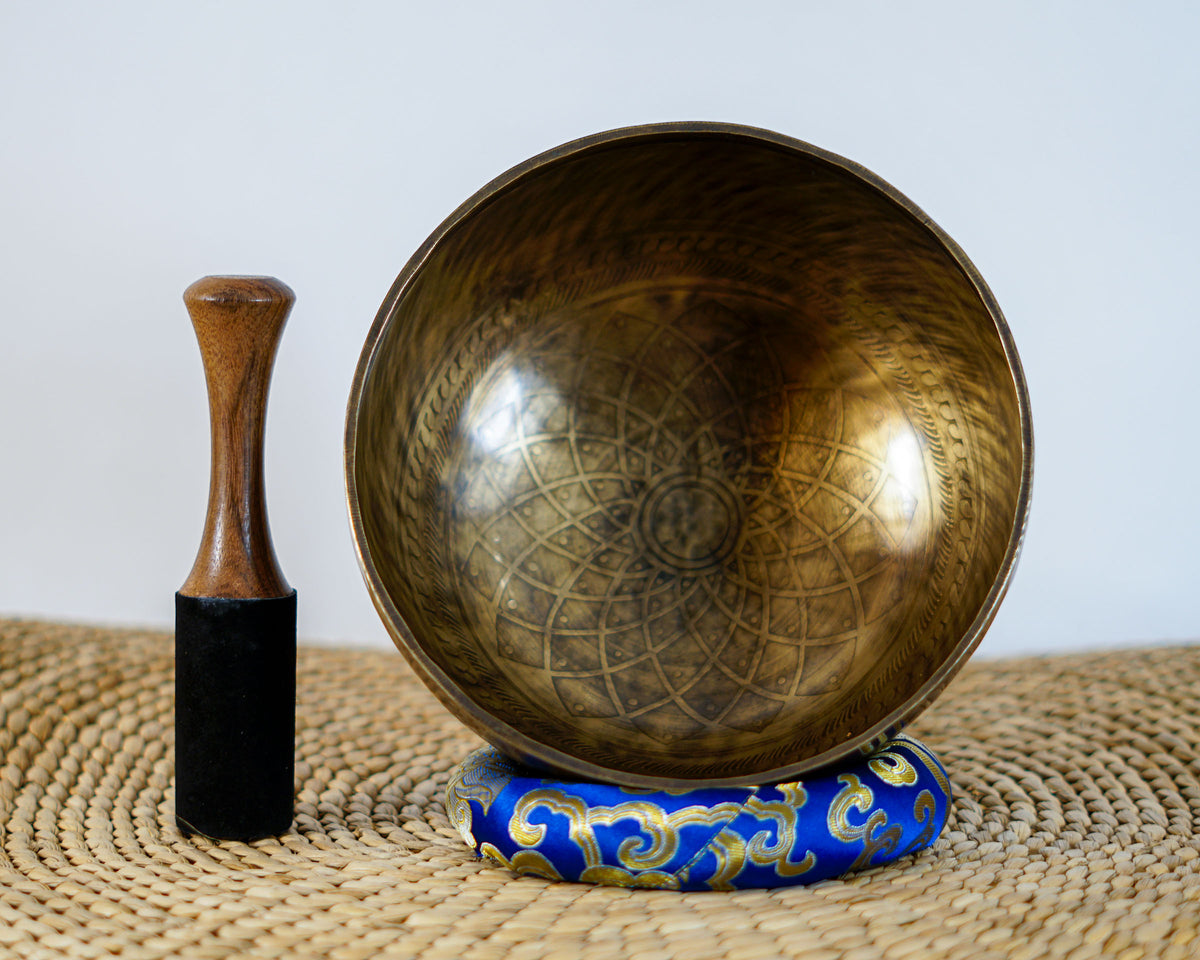 Tibetan singing bowl displaying a cosmic geometric pattern inside, with a wooden mallet and blue brocade cushion.