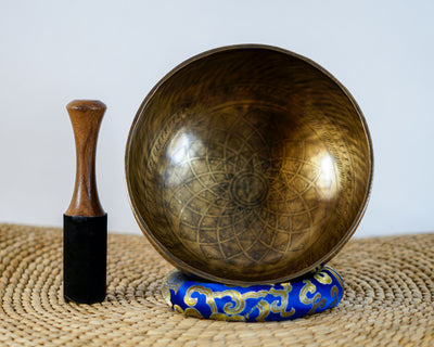 Tibetan singing bowl displaying a cosmic geometric pattern inside, with a wooden mallet and blue brocade cushion.