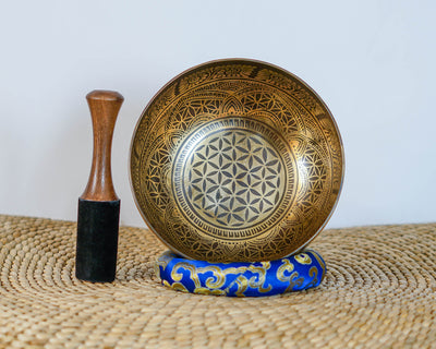 Flower of Life engraved Tibetan singing bowl displayed upright with a wooden mallet and blue brocade cushion.