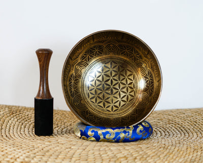 Engraved Tibetan singing bowl featuring a Flower of Life design, photographed upright with a wooden mallet and blue brocade cushion.