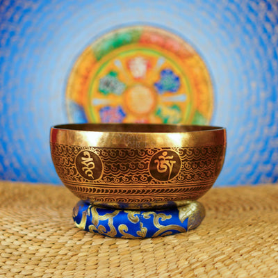 Handcrafted Flower of Life singing bowl from Nepal with sacred geometry engraving, Tibetan-style motifs, bronze-gold finish and blue cushion.