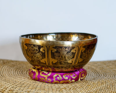 Golden Tibetan singing bowl featuring engraved Buddhist symbols and decorative patterns, displayed on a pink brocade cushion.