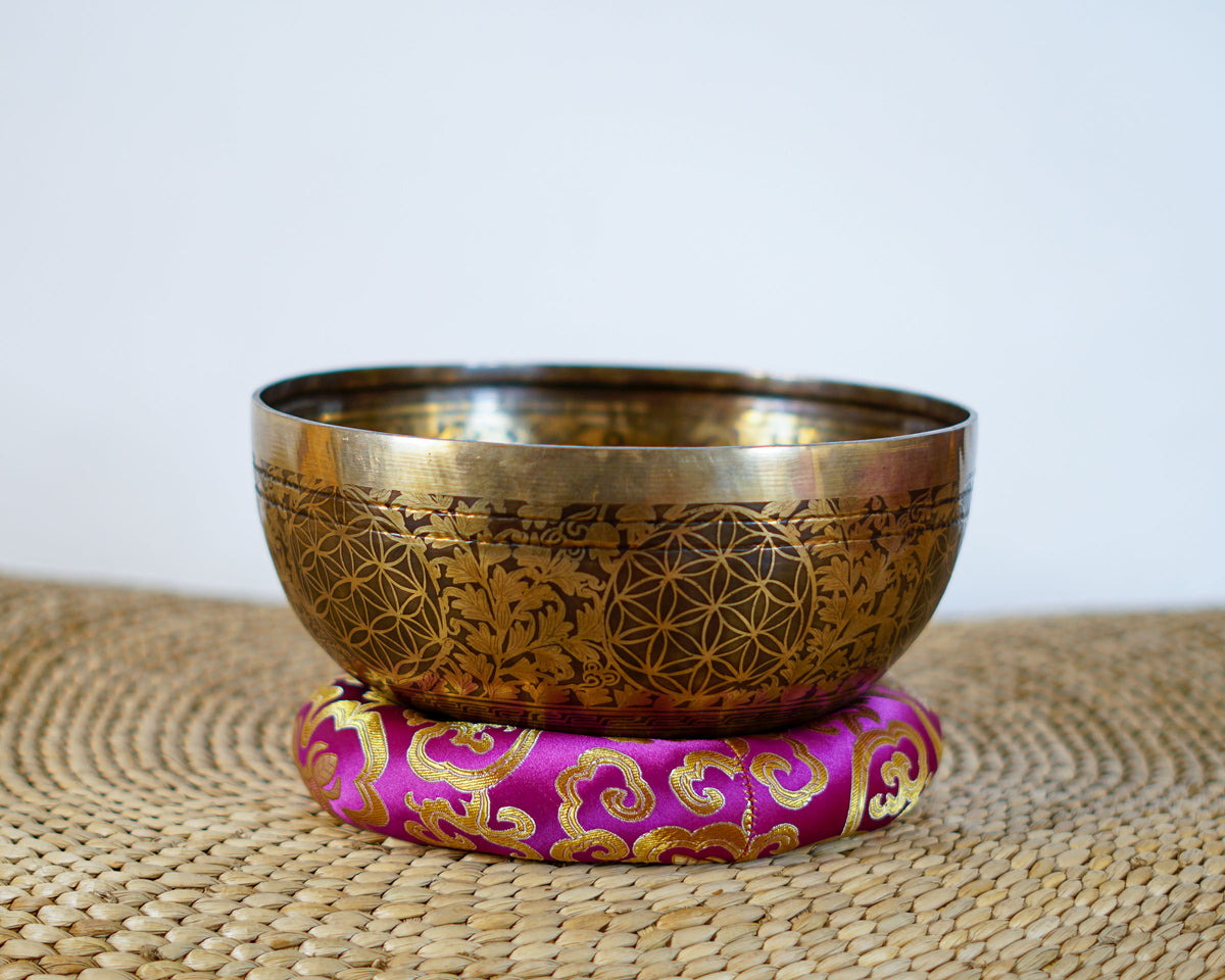 Golden and bronze Chengresi singing bowl featuring detailed floral engravings, displayed on a pink brocade cushion.