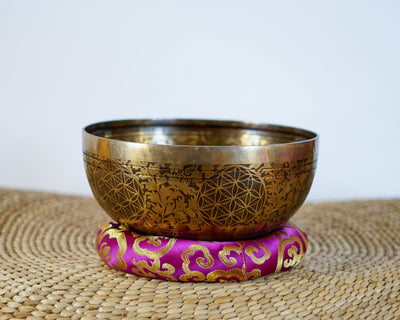 Golden and bronze Chengresi singing bowl featuring detailed floral engravings, displayed on a pink brocade cushion.