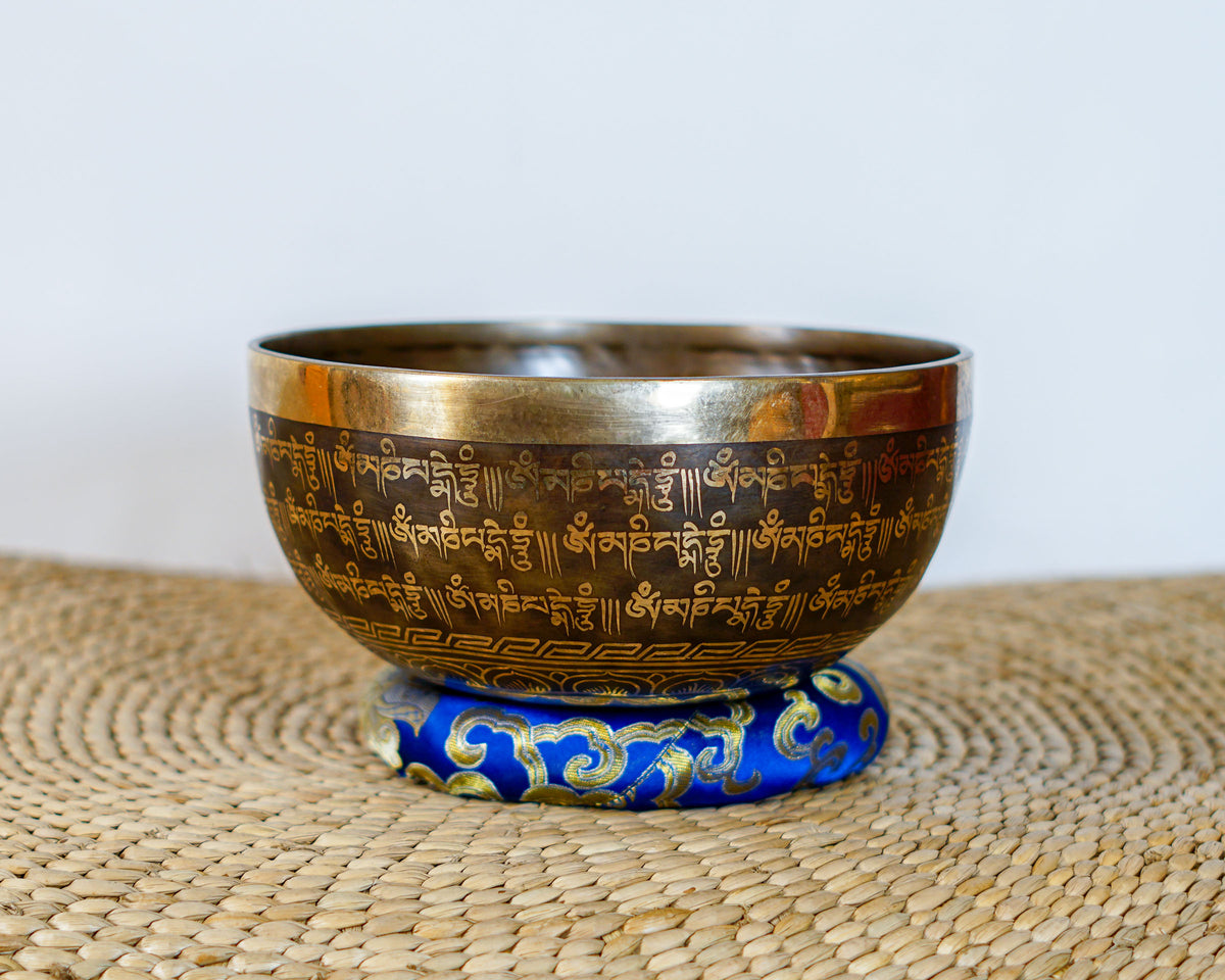 Exterior side view of a dark antique bronze Tibetan singing bowl engraved with golden Tibetan script, placed on a blue brocade cushion.