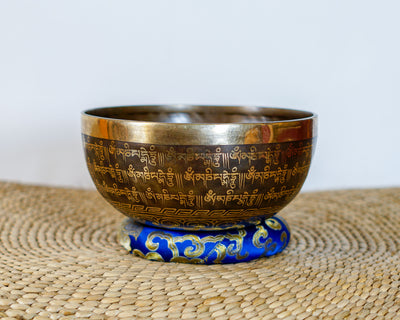 Exterior side view of a dark antique bronze Tibetan singing bowl engraved with golden Tibetan script, placed on a blue brocade cushion.