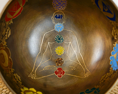 Close up inside a dark bronze Tibetan singing bowl showing a meditating yogi figure with seven hand-painted colored chakra symbols.