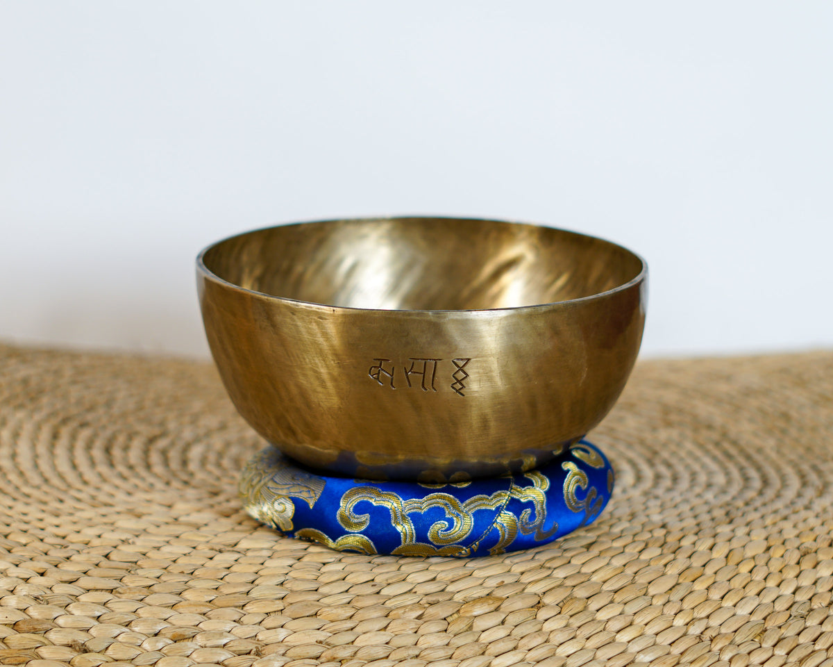 Side view of a warm golden hand-hammered Kasa Moon singing bowl showing the engraved Devanagari script on a blue brocade cushion.