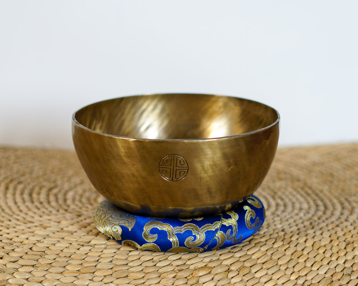 Side view of a warm golden hand-hammered Kasa Moon singing bowl showing the engraved Devanagari script on a blue brocade cushion.