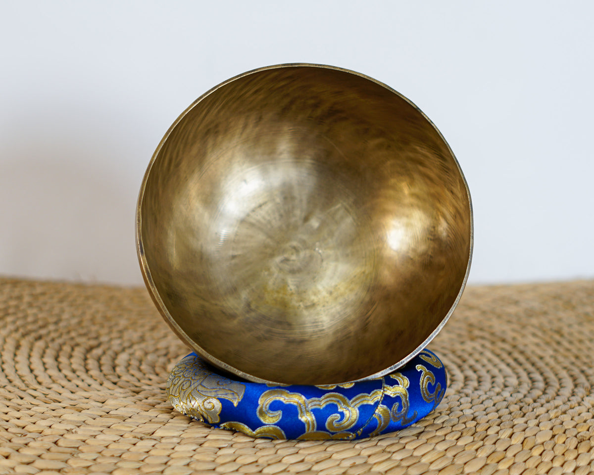 Top-down view into the smooth golden interior of a hand-hammered Kasa Moon Tibetan singing bowl resting on a blue brocade cushion.