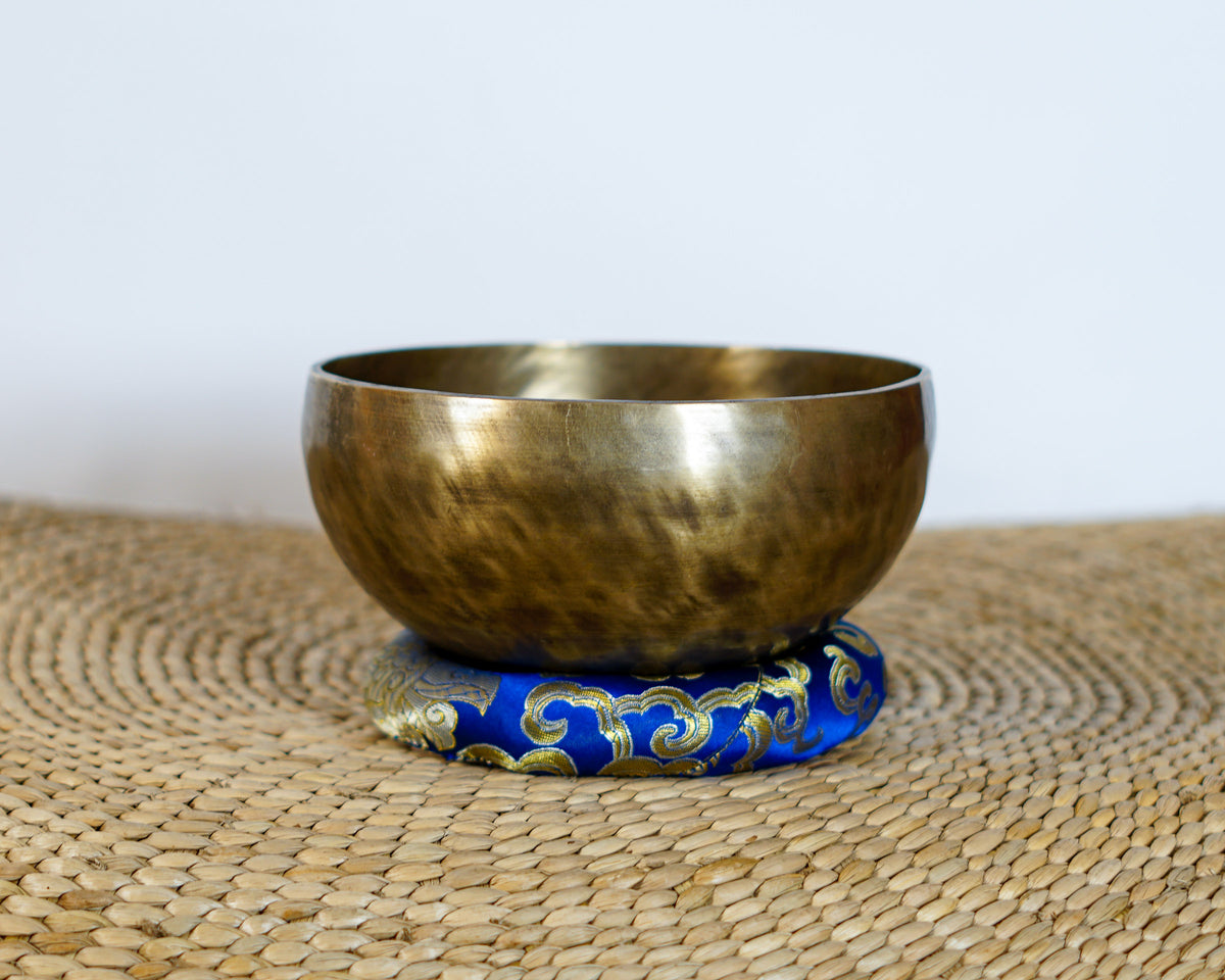 Side view of a hand-hammered antique bronze Tibetan singing bowl displayed on a blue brocade cushion.