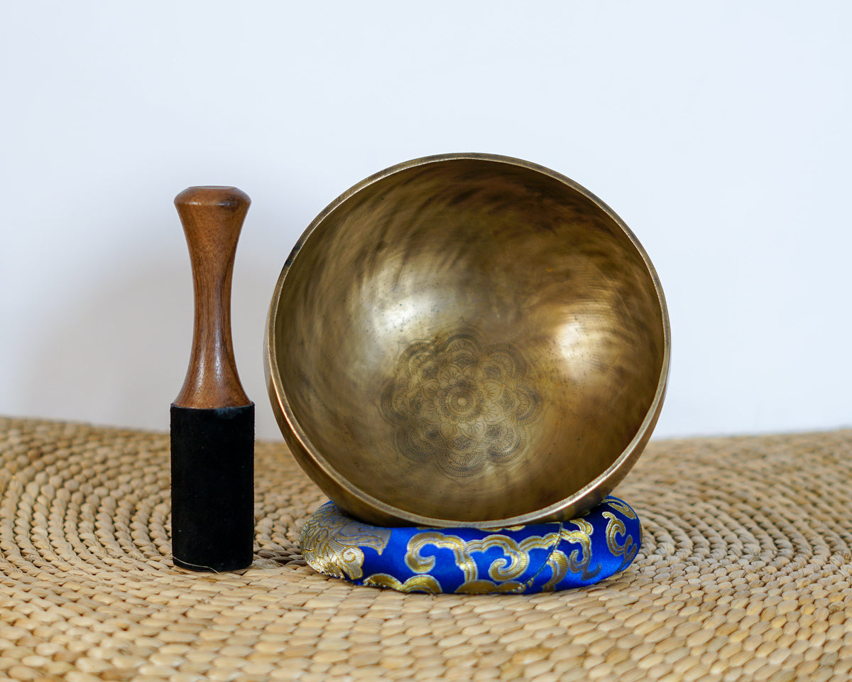 Top-angled view of an antique bronze Tibetan singing bowl showing the interior lotus flower mandala engraving, shown with a wooden mallet striker and blue cushion.